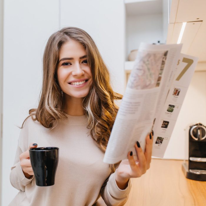 Attractive girl with light brown hair holding magazine and laughing in her room. Indoor photo of cute brunette woman with cup of tea and newspaper..